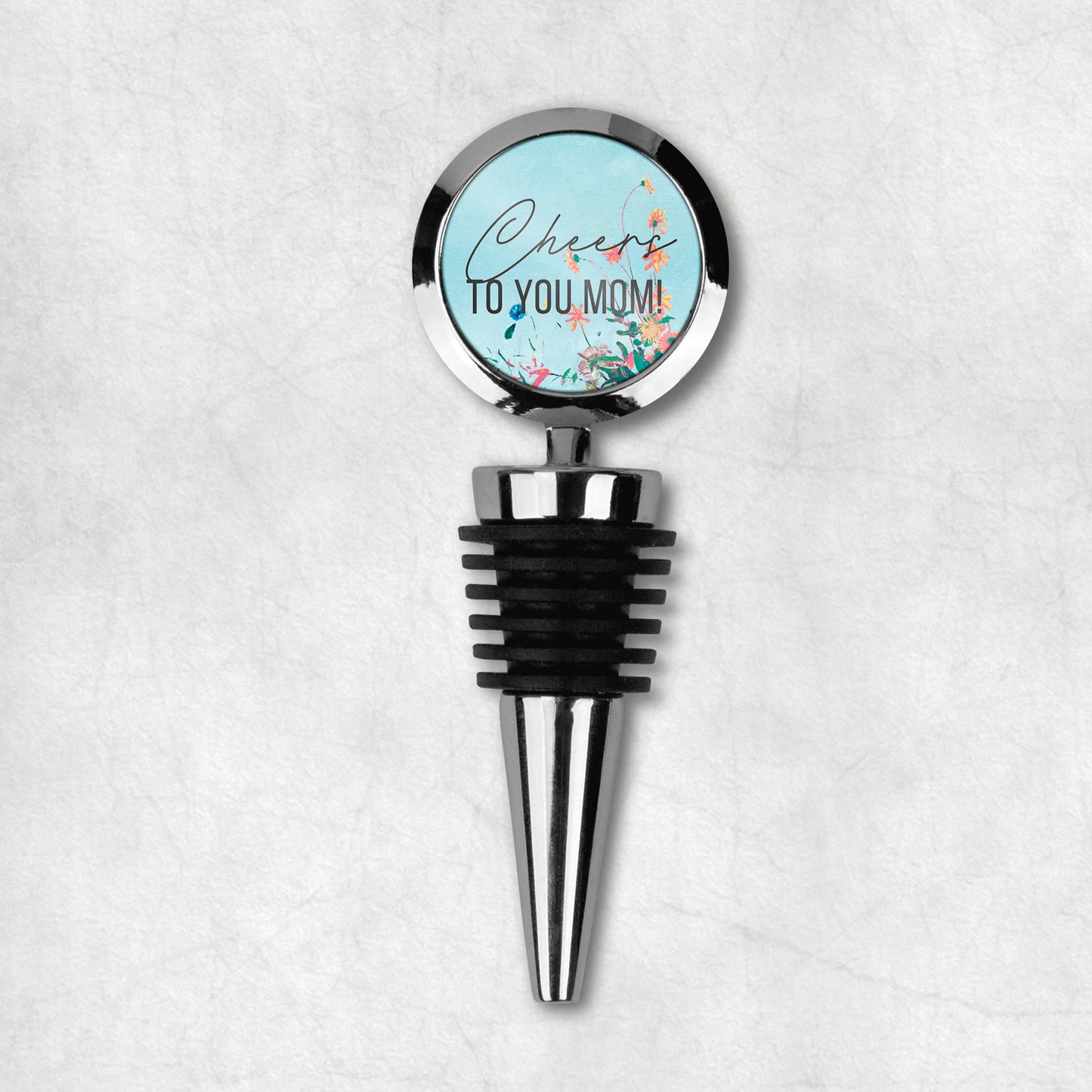 A wine stopper with a round top with 'Cheers to you mom' text and a floral design, predominantly in black, blue, and green colors, with a metal stem and rubber stopper.