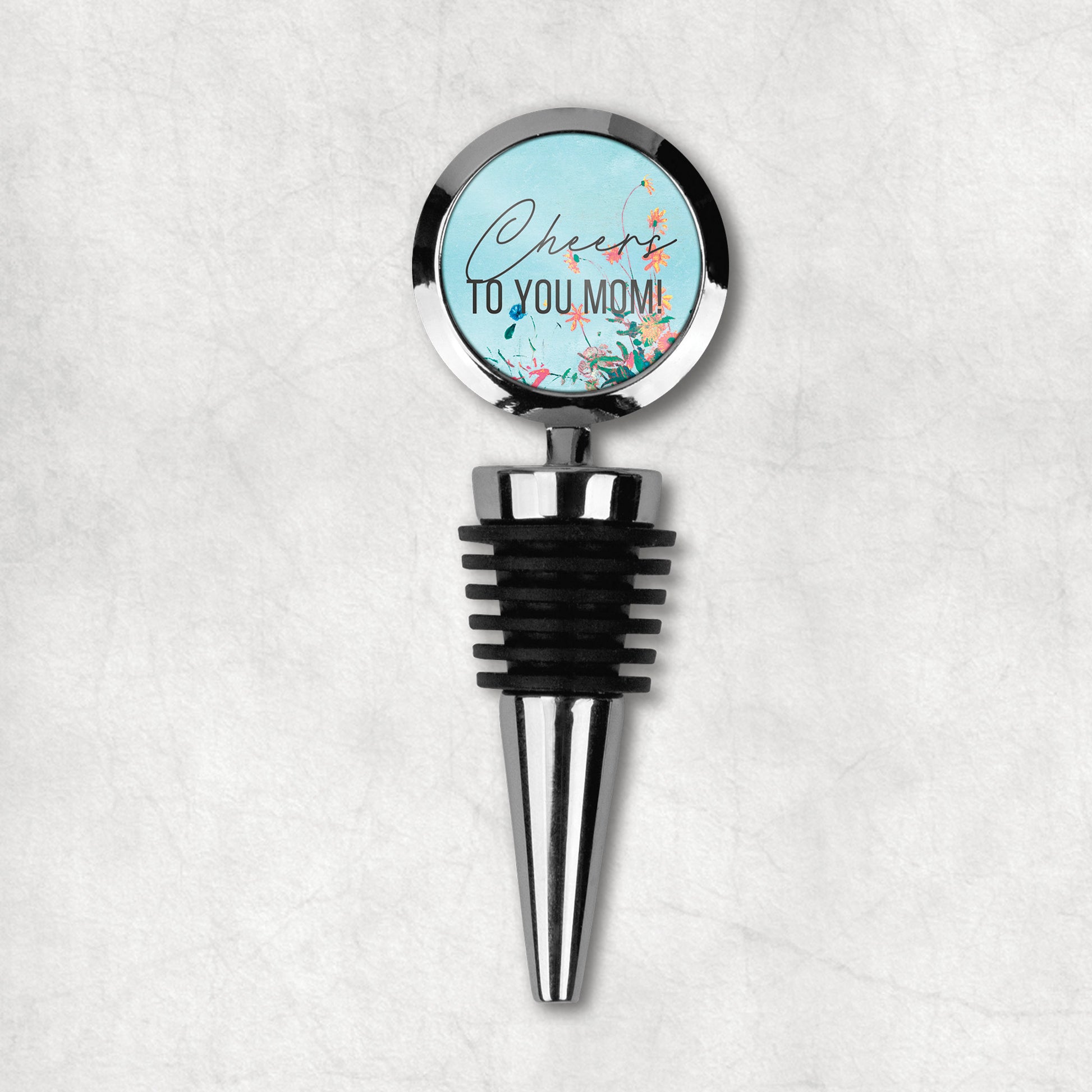 A wine stopper with a round top with 'Cheers to you mom' text and a floral design, predominantly in black, blue, and green colors, with a metal stem and rubber stopper.