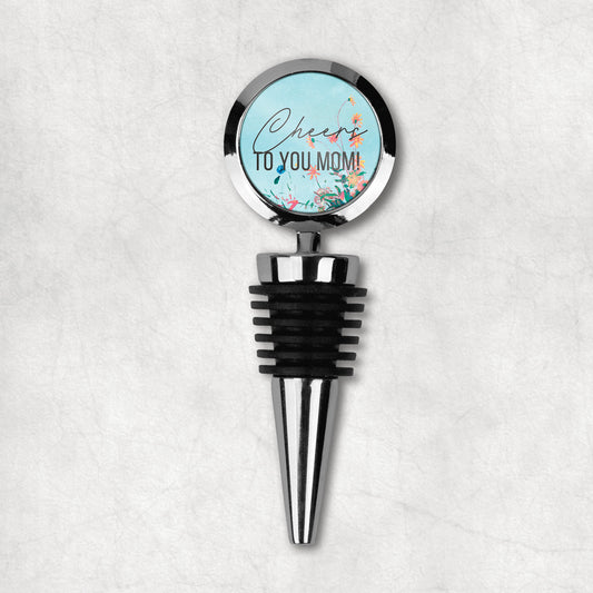 A wine stopper with a round top with 'Cheers to you mom' text and a floral design, predominantly in black, blue, and green colors, with a metal stem and rubber stopper.