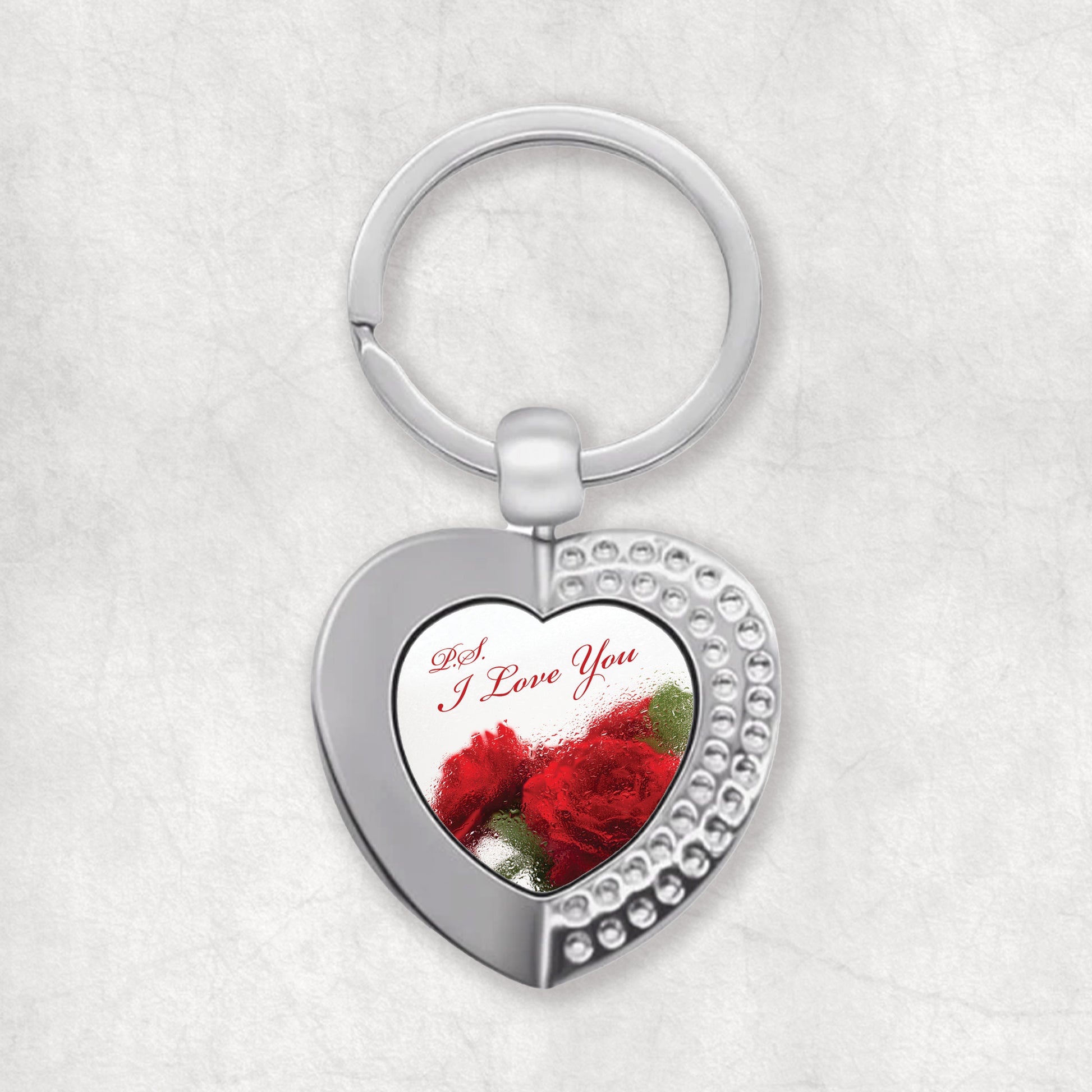 A heart-shaped keyring with a single-sided print of the phrase 'P.S. I Love You' surrounded by red decorative elements, attached to a silver-colored ring.