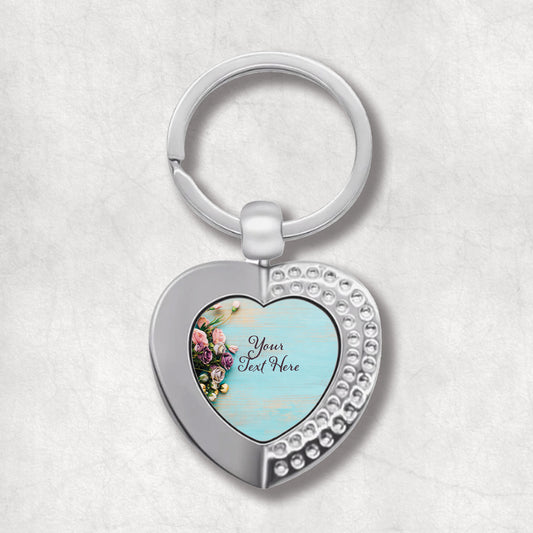 An elegant heart-shaped keyring with a floral design and a space for personalised text, predominantly silver and blue.