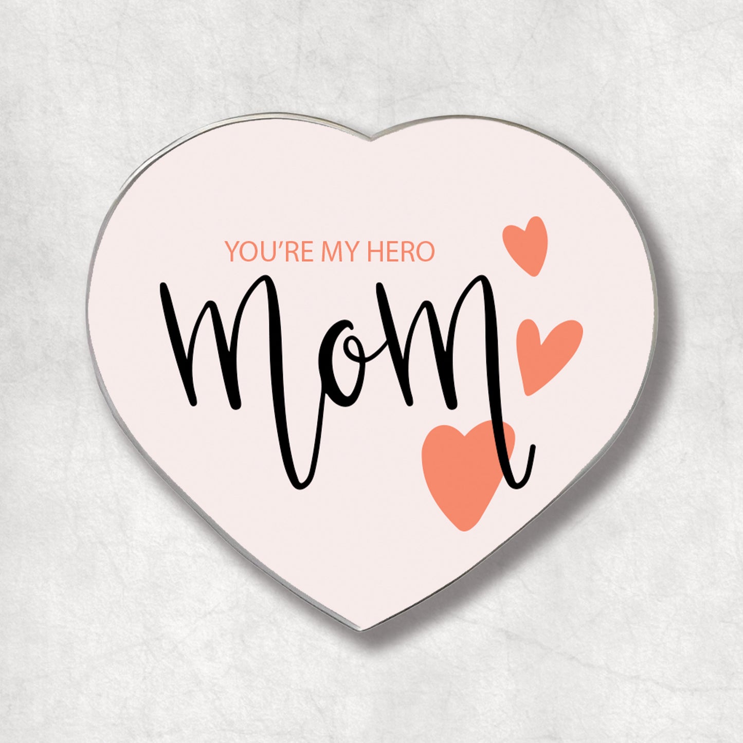 A heart-shaped metal magnet with the text 'YOU'RE MY HERO' and 'mom' in curly black font, accompanied by two pink hearts.