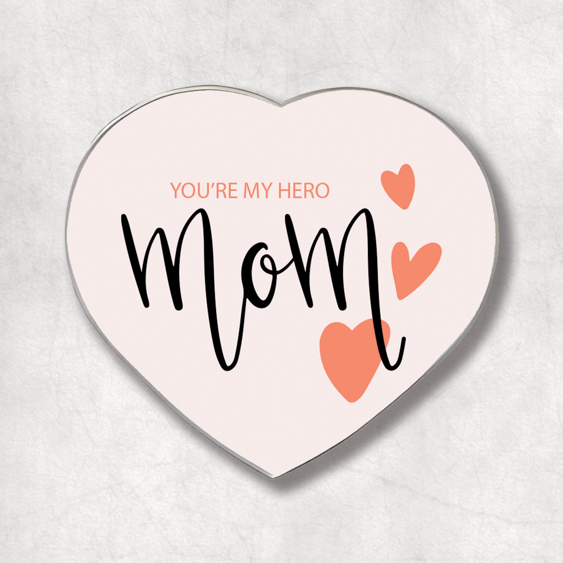 A heart-shaped metal magnet with the text 'YOU'RE MY HERO' and 'mom' in curly black font, accompanied by two pink hearts.
