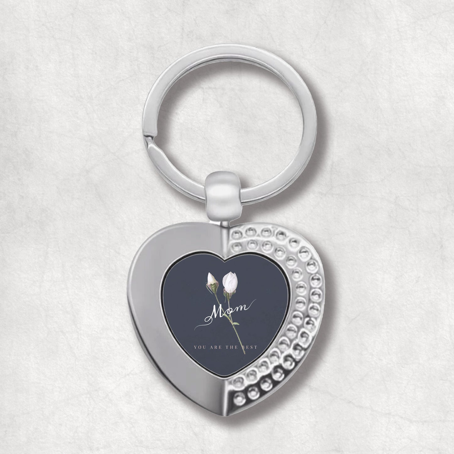 An elegant heart-shaped keyring with a printed message 'Mom you are the best' with white roses on a dark background.