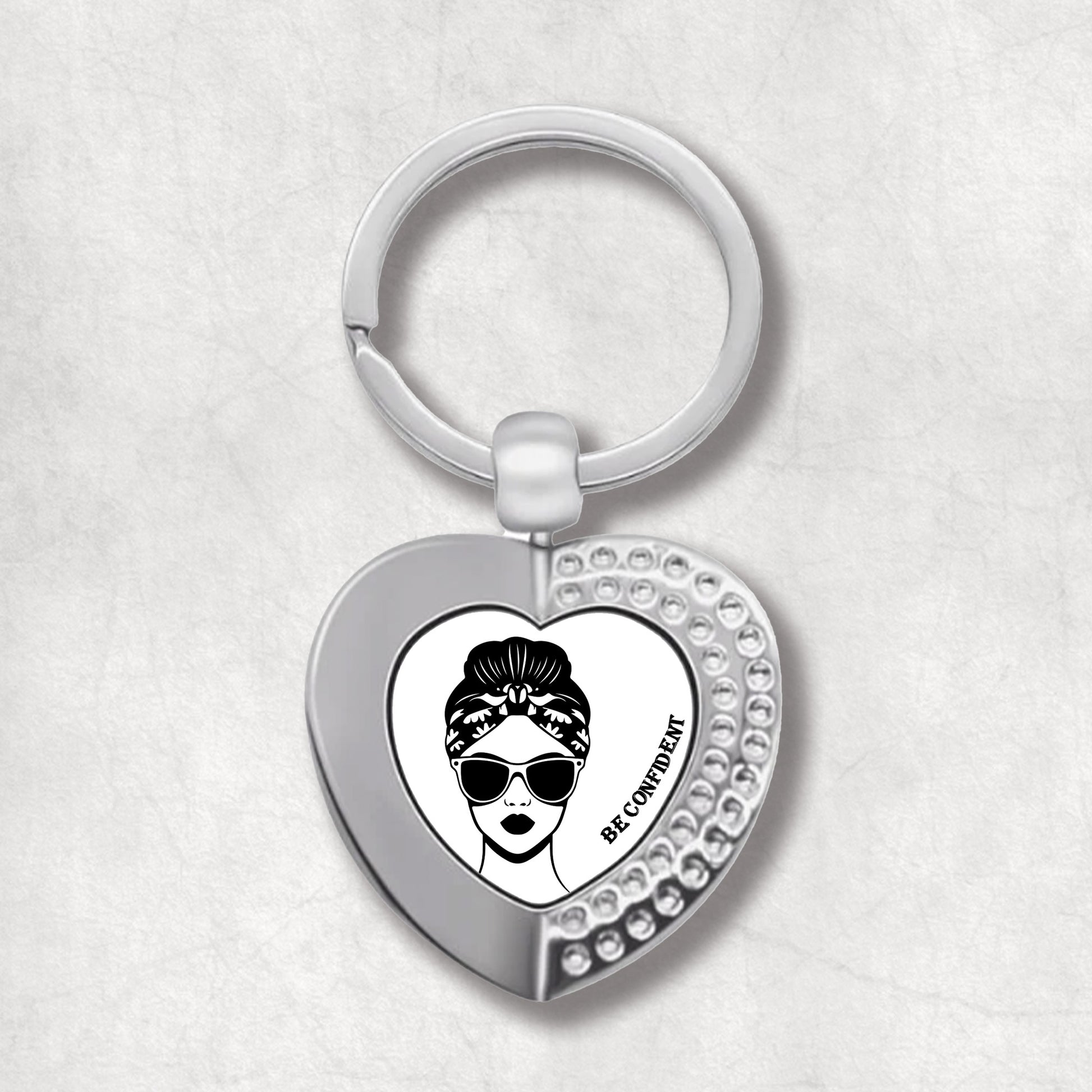 Metal heart-shaped keychain with "Be Confident" text and a woman's face wearing sunglasses graphic, on a white background.