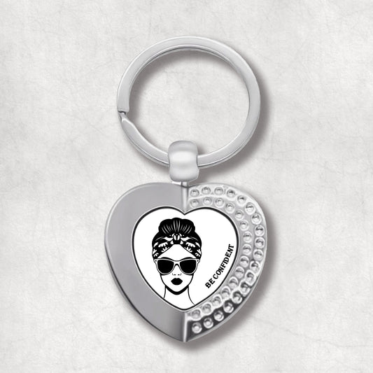 Metal heart-shaped keychain with "Be Confident" text and a woman's face wearing sunglasses graphic, on a white background.