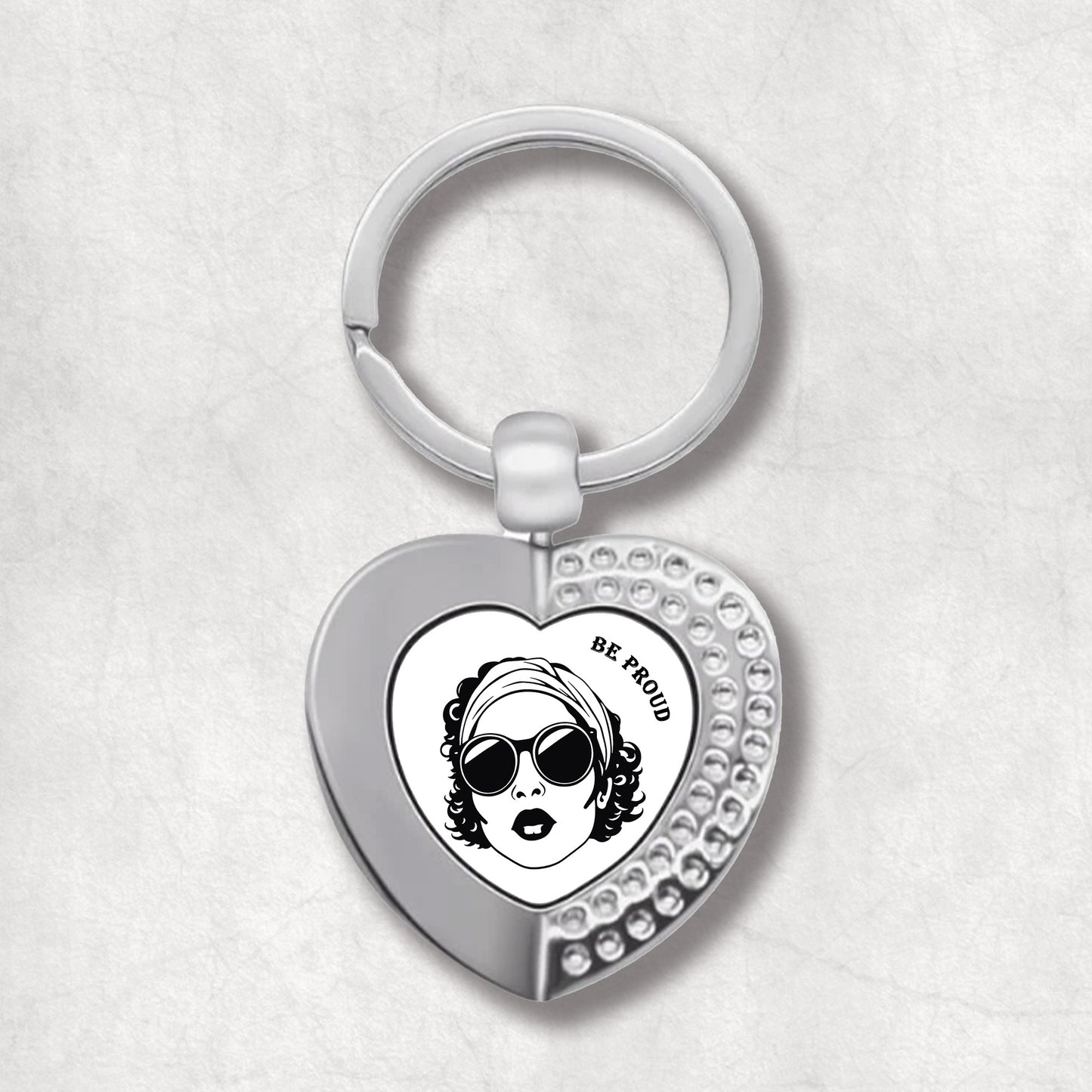 Metal heart-shaped keychain with "Be Proud" text and a woman's face graphic on a white background.