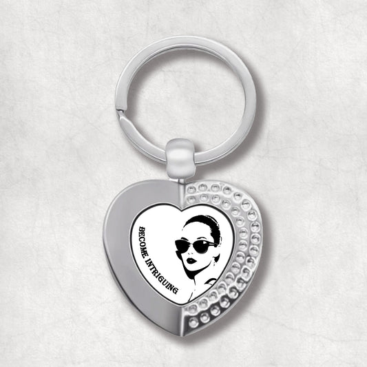 Metal heart-shaped keychain with 'Become Intriguing' text and a woman's face graphic on a white background.  