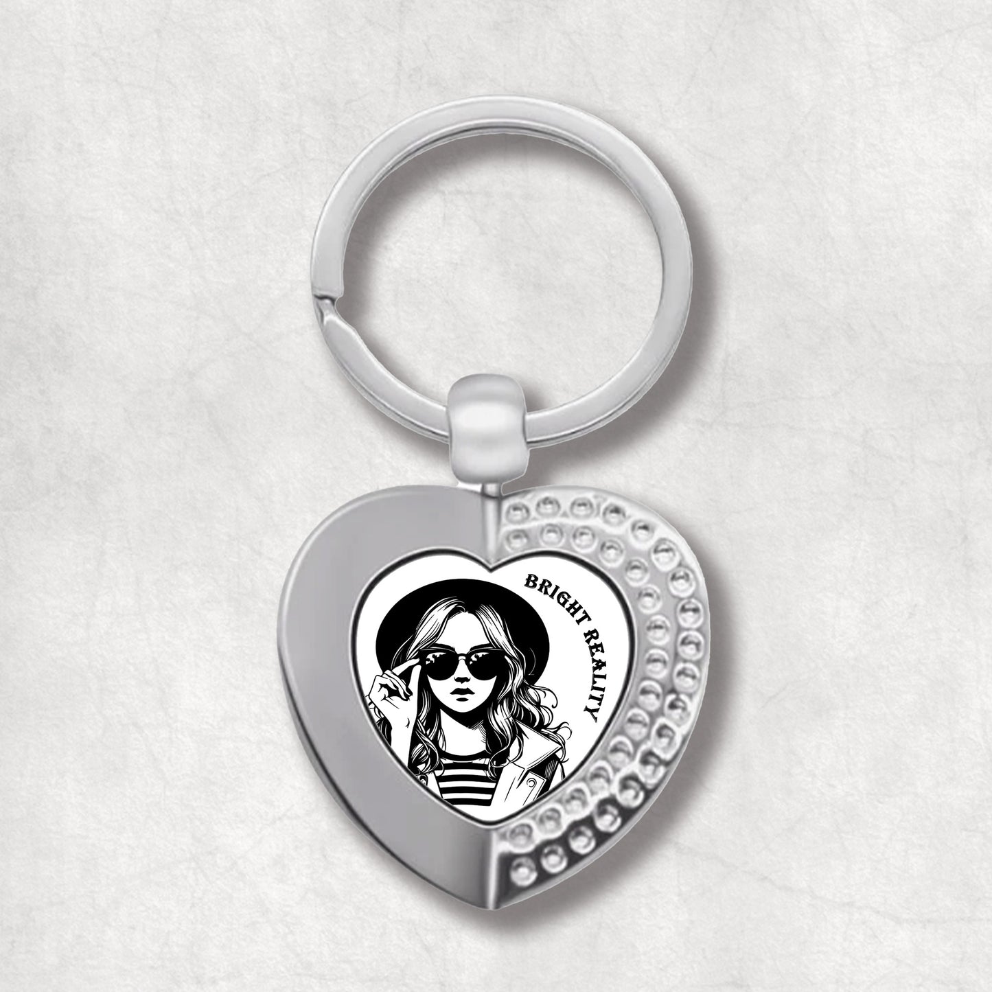 Metal heart-shaped keychain with a black and white illustration of a woman wearing sunglasses, and text 'bright reality' on a white background.