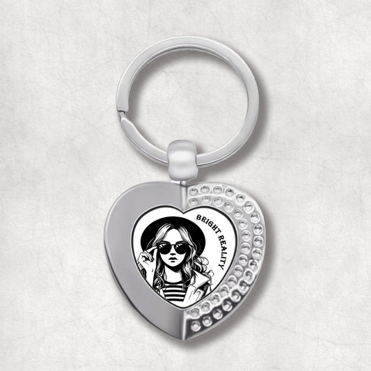 Metal heart-shaped keychain with a black and white illustration of a woman wearing sunglasses, and text 'bright reality' on a white background.