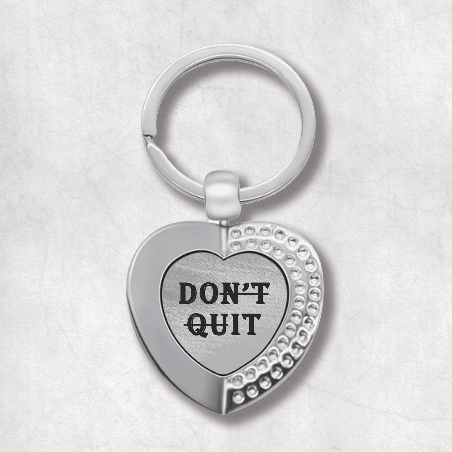 Metal heart-shaped keychain with 'don't quit' text written on a grey patterned background.