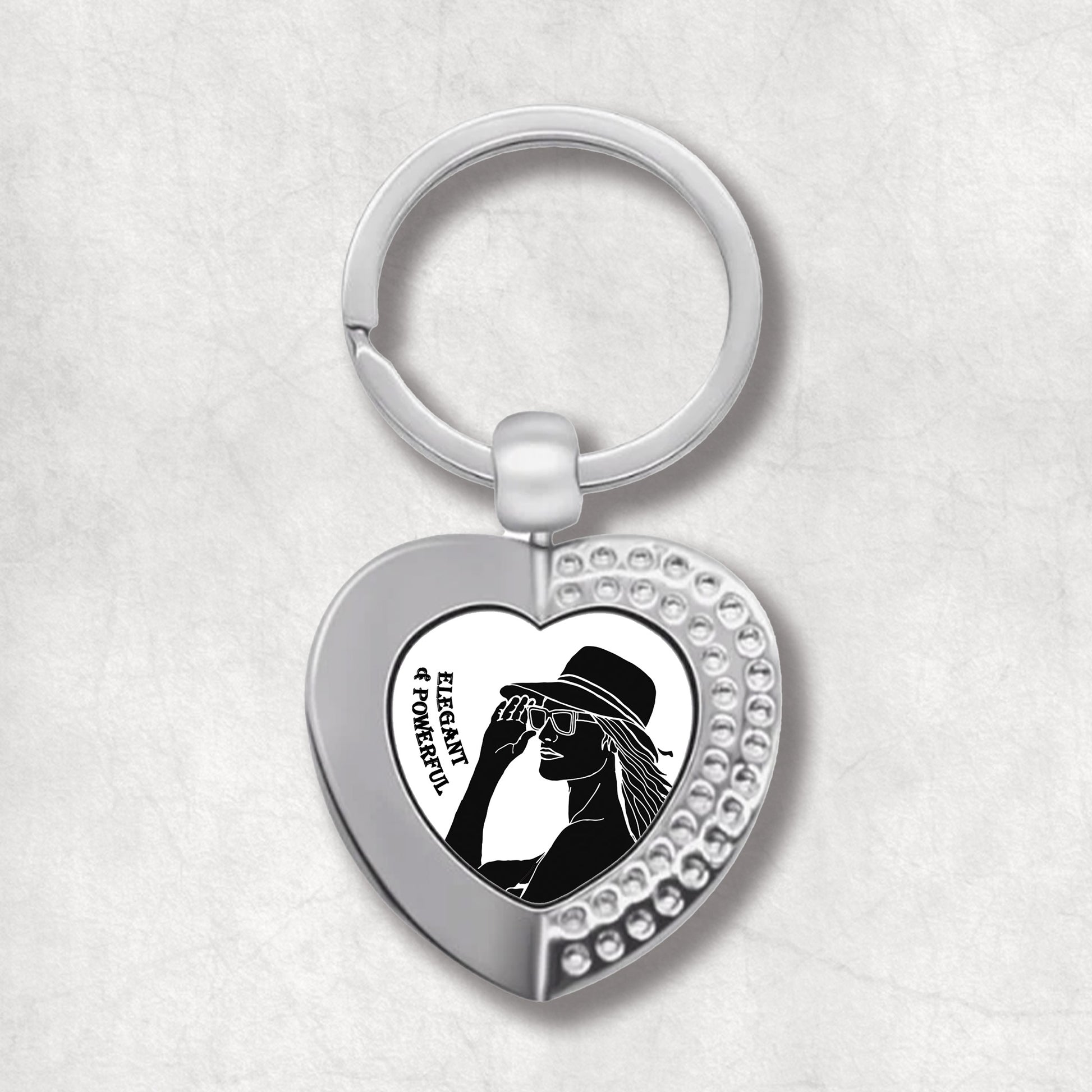 Metal heart-shaped keychain with a silhouette illustration of a woman, and text 'Elegant & Powerful' written on a white background.