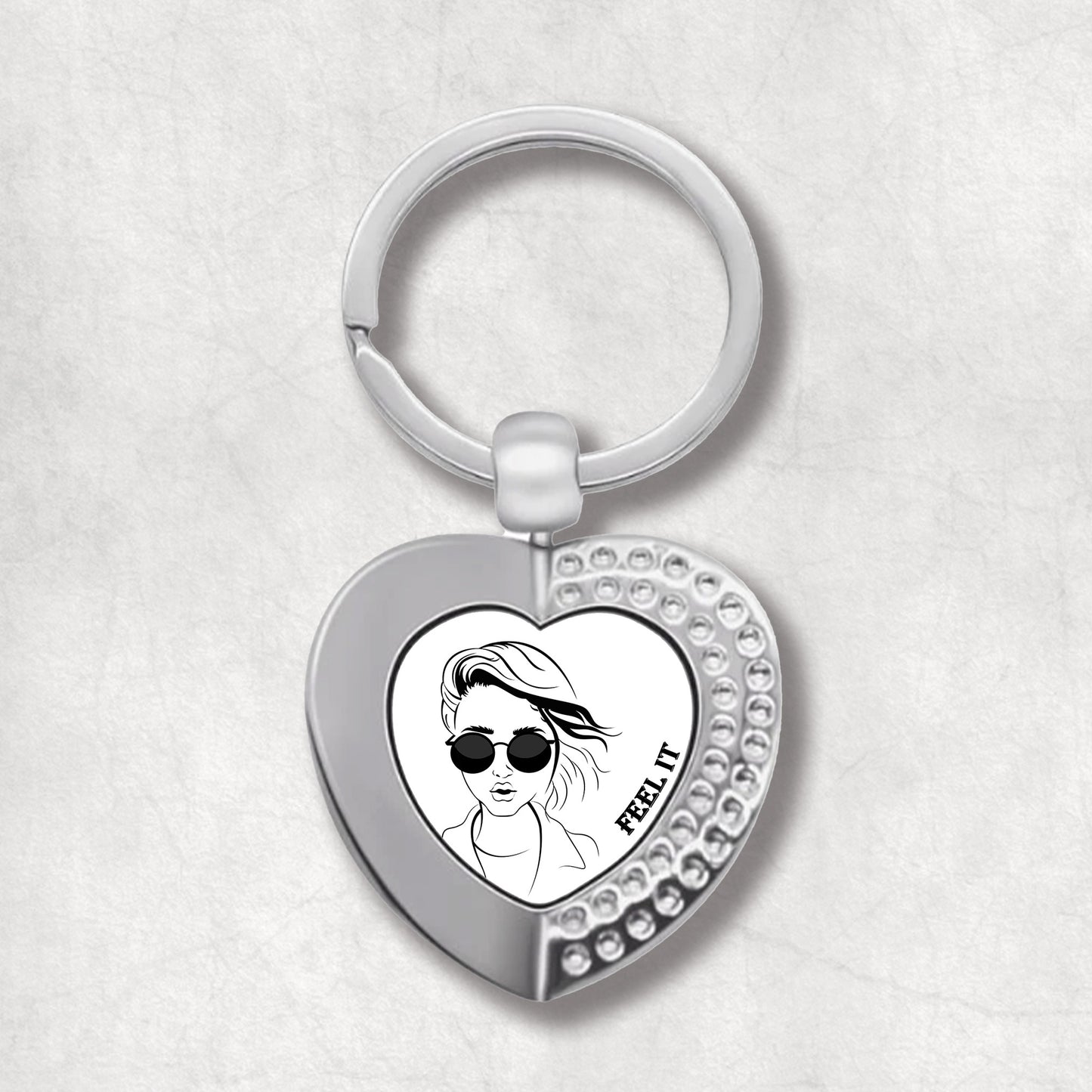 Metal heart-shaped keychain with a design of a woman wearing sunglasses and the text 'feel it' written on a white background.