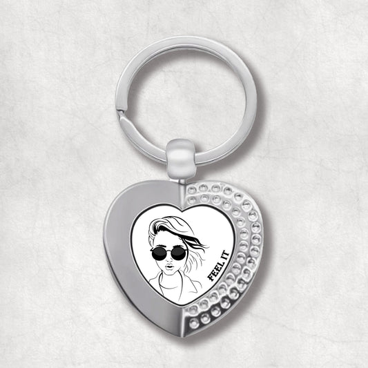 Metal heart-shaped keychain with a design of a woman wearing sunglasses and the text 'feel it' written on a white background.