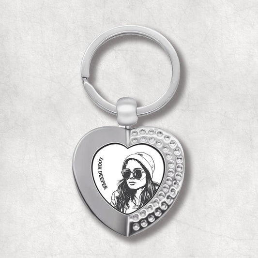 Metal heart-shaped keychain with a woman's face wearing a hat, sunglasses and 'Look Deeper' text written on a light background.