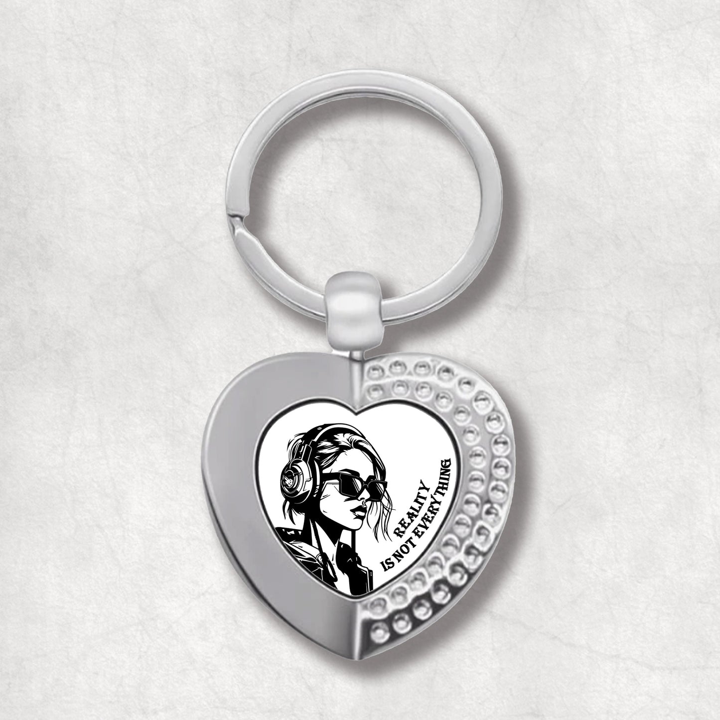 Metal heart-shaped keychain with an illustration of a woman wearing sunglasses, headphones and 'reality is not everything' text written on a white background.