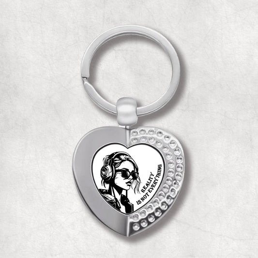 Metal heart-shaped keychain with an illustration of a woman wearing sunglasses, headphones and 'reality is not everything' text written on a white background.