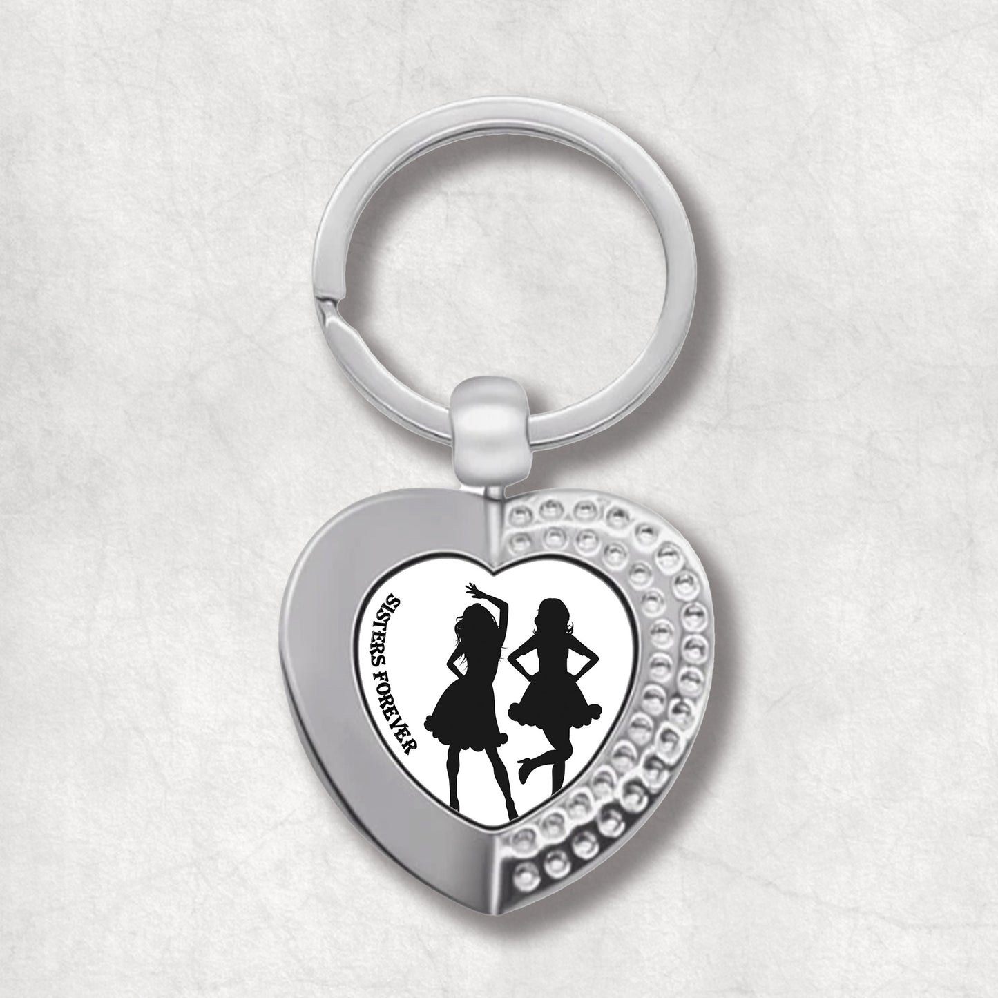 Metal heart-shaped keychain with silhouettes of dancing women and 'Sisters Forever' text written on a white background.