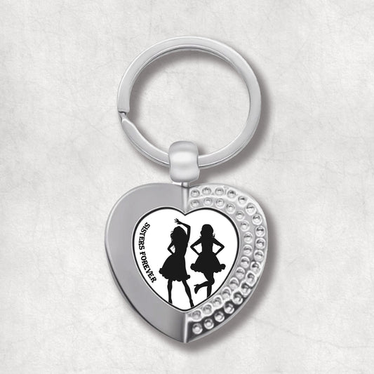 Metal heart-shaped keychain with silhouettes of dancing women and 'Sisters Forever' text written on a white background.