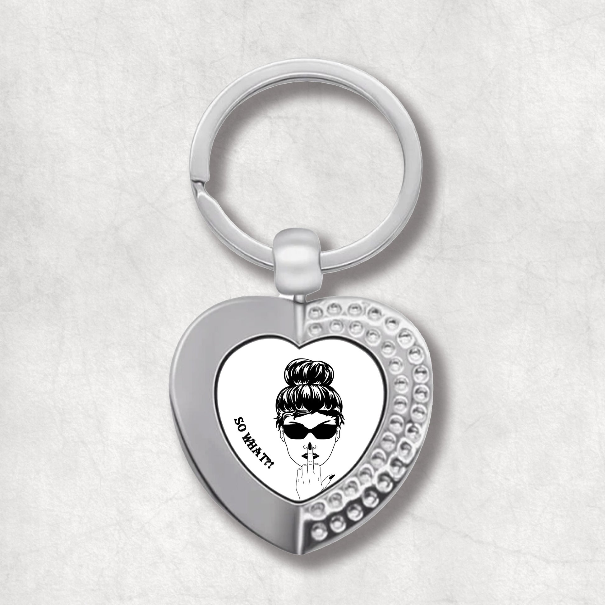 Metal heart-shaped keychain with an illustration of a woman's face, wearing sunglasses, showing middle finger 'so what?' text written on a white background.