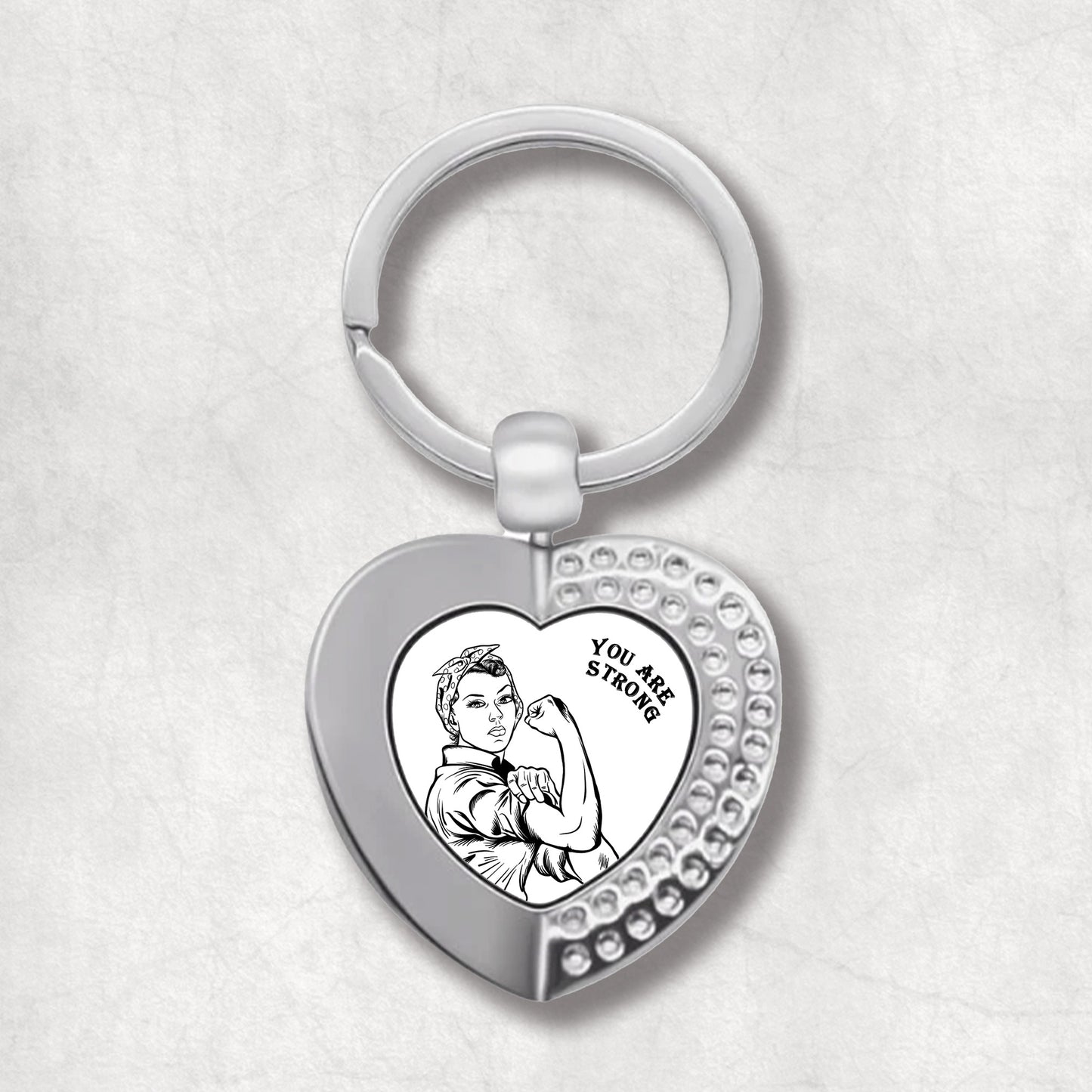 Metal heart-shaped keychain with a black and white, vintage-style illustration of a strong woman, and 'You Are Strong' text written on a white background.