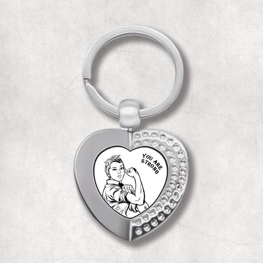 Metal heart-shaped keychain with a black and white, vintage-style illustration of a strong woman, and 'You Are Strong' text written on a white background.